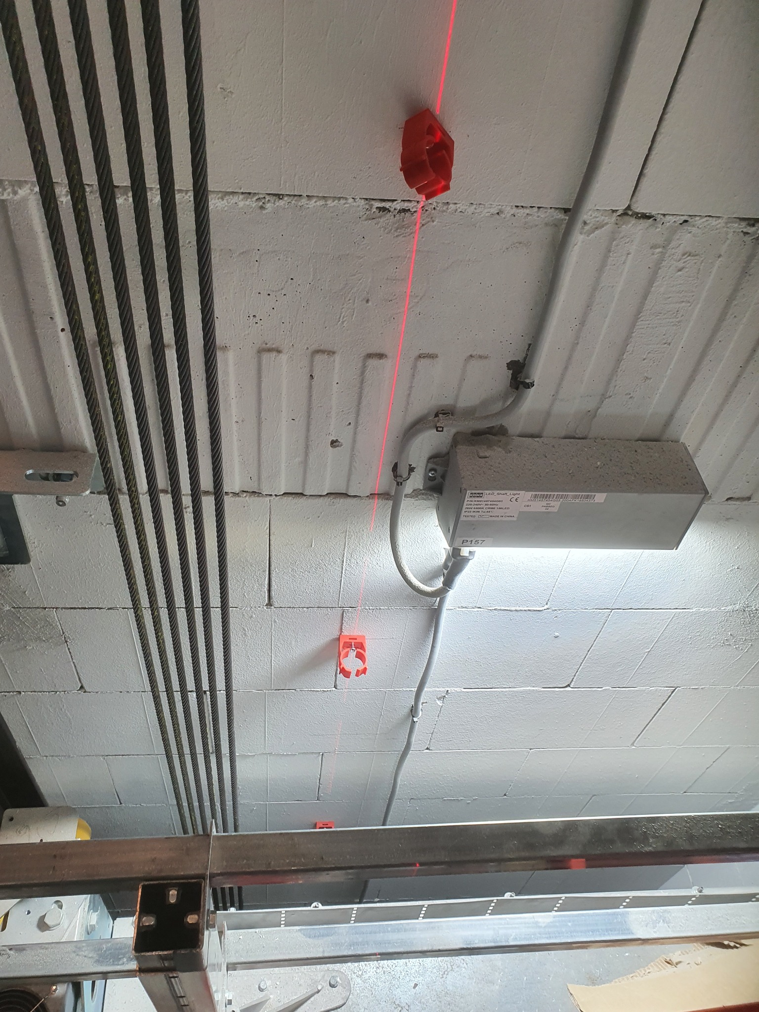 Laserowy pomiar pionu w szybie windy, widoczne kable, oświetlenie i elementy konstrukcyjne. Czerwona linia lasera wyznacza pion.