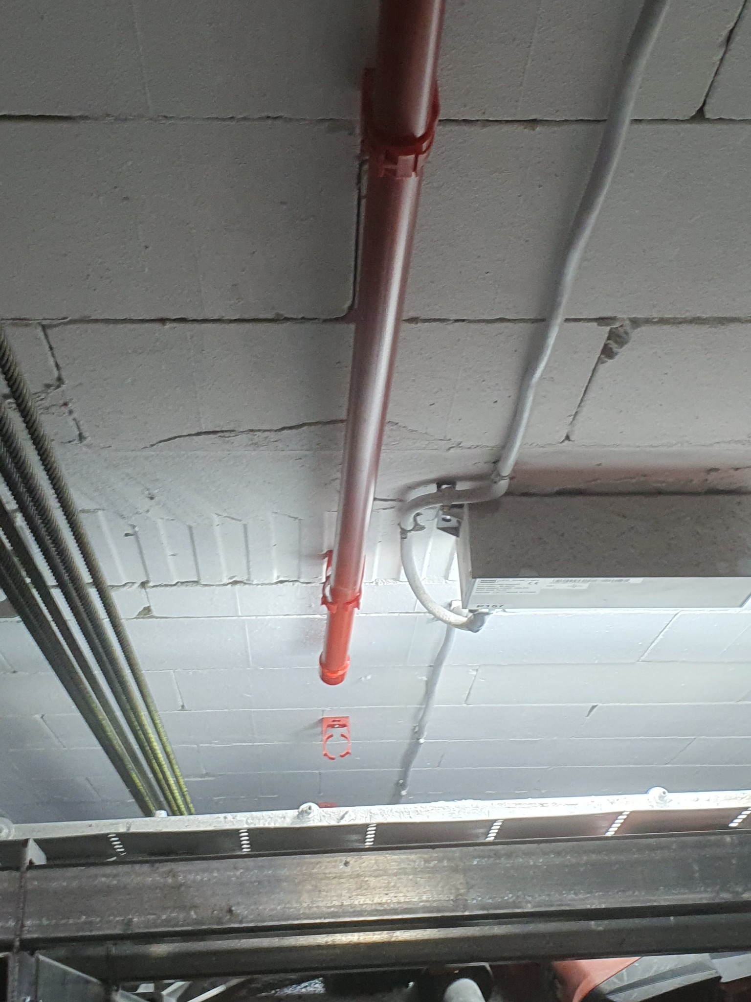 Instalacja rur i kabli na suficie z bloków betonowych, widok z dołu. Czerwona rura pionowo, szare kable poziomo. Metalowa konstrukcja w dolnej części kadru.