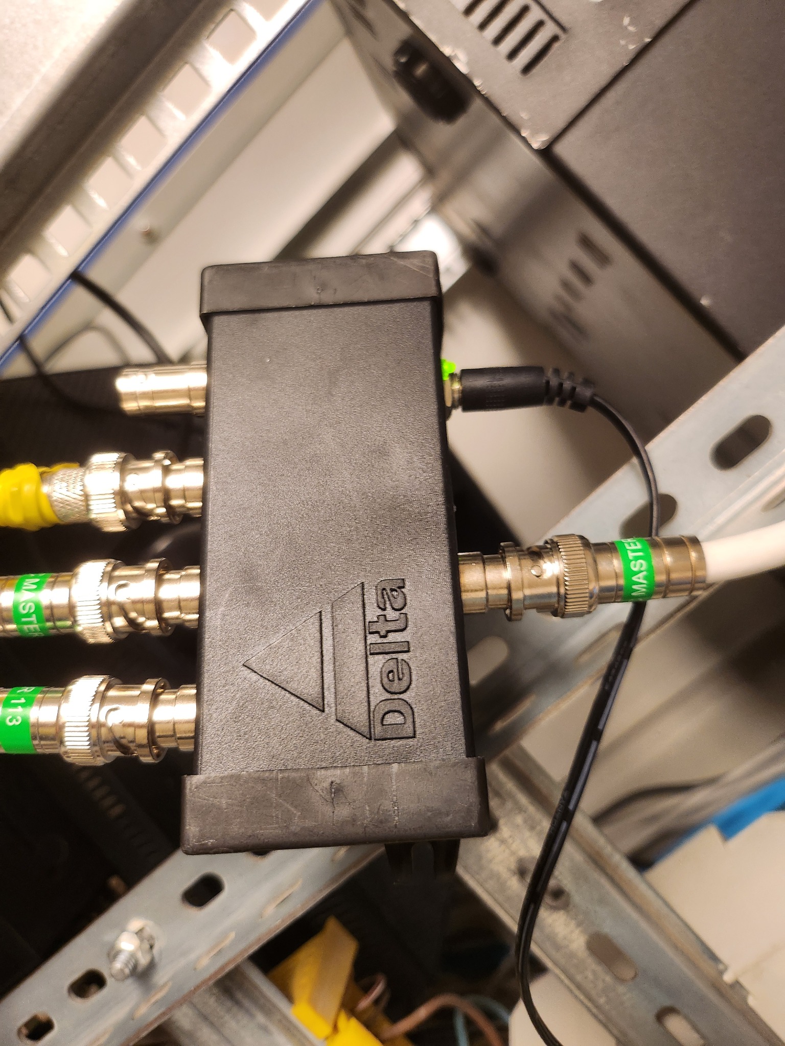 Czarny rozdzielacz Delta z podłączonymi kablami BNC i zasilaniem w szafie rack, widoczne oznaczenia 'MASTER' na kablach. Element instalacji alarmowej lub monitoringu.