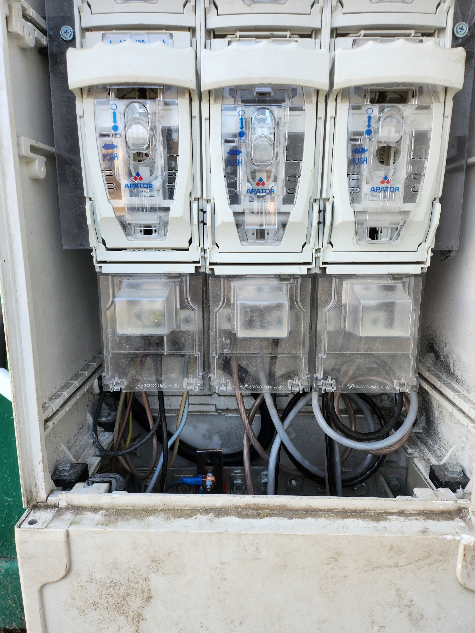Otwarta skrzynka z trzema licznikami energii elektrycznej marki Apator, widoczne przewody i elementy montażowe wewnątrz. Obudowa lekko zabrudzona.
