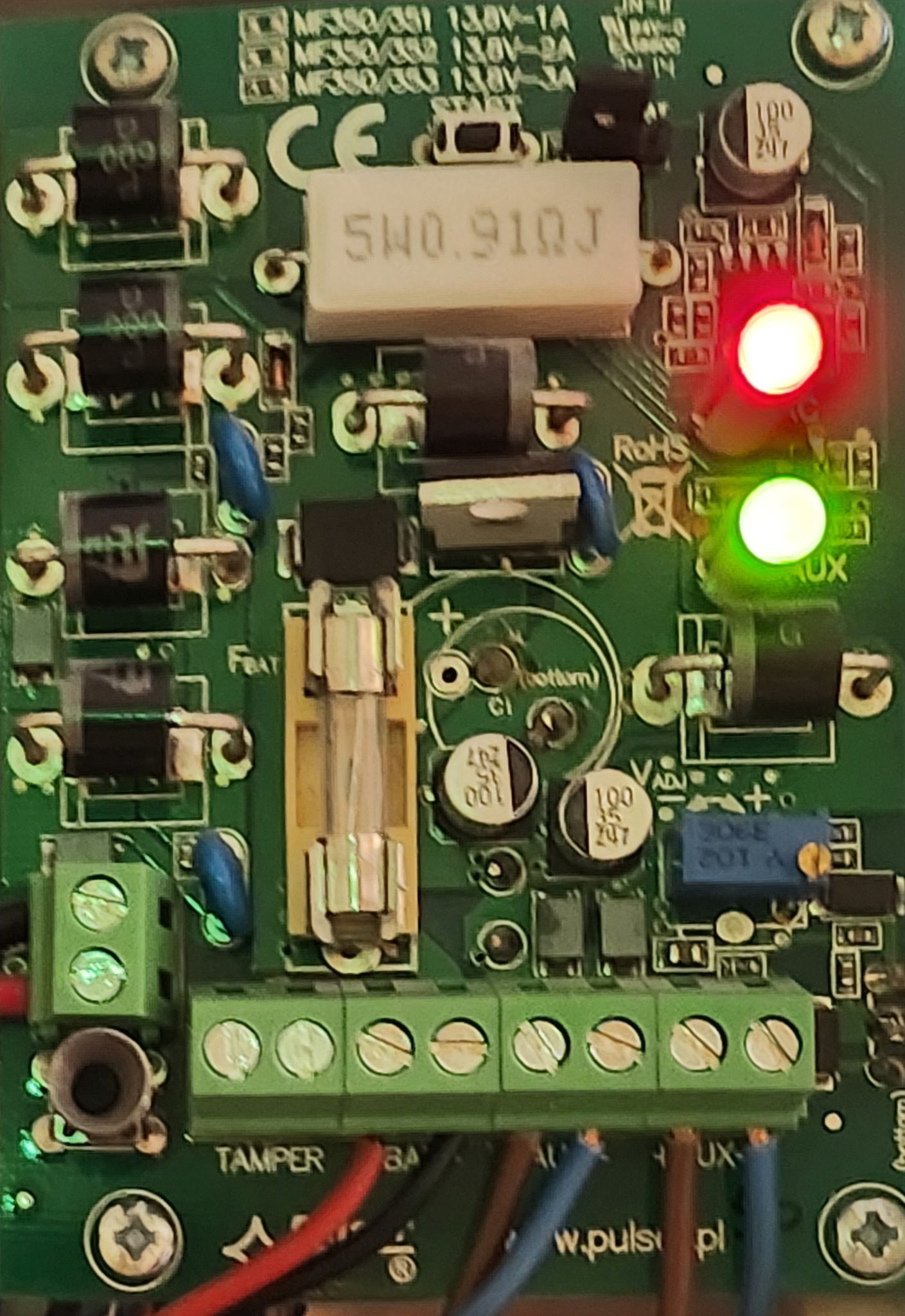 Zbliżenie na zieloną płytkę drukowaną z elementami elektronicznymi, w tym diodami LED (czerwona i zielona), rezystorami i złączami. Widoczne kable podłączone do listwy zaciskowej.