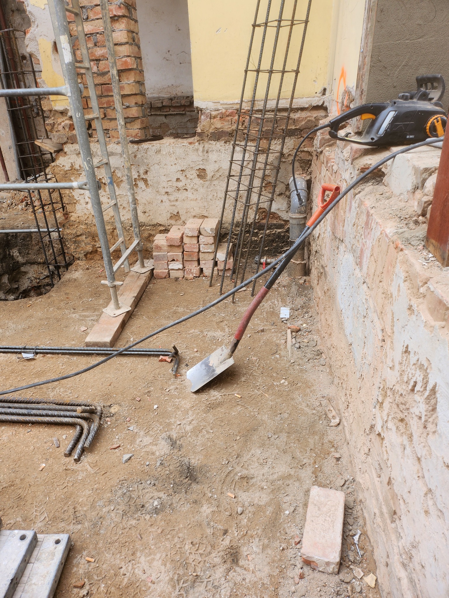 Przygotowania do fundamentów: wykop, zbrojenie, łopata i piła elektryczna na tle ceglanej ściany. Widoczne ślady prac ziemnych i budowlanych.