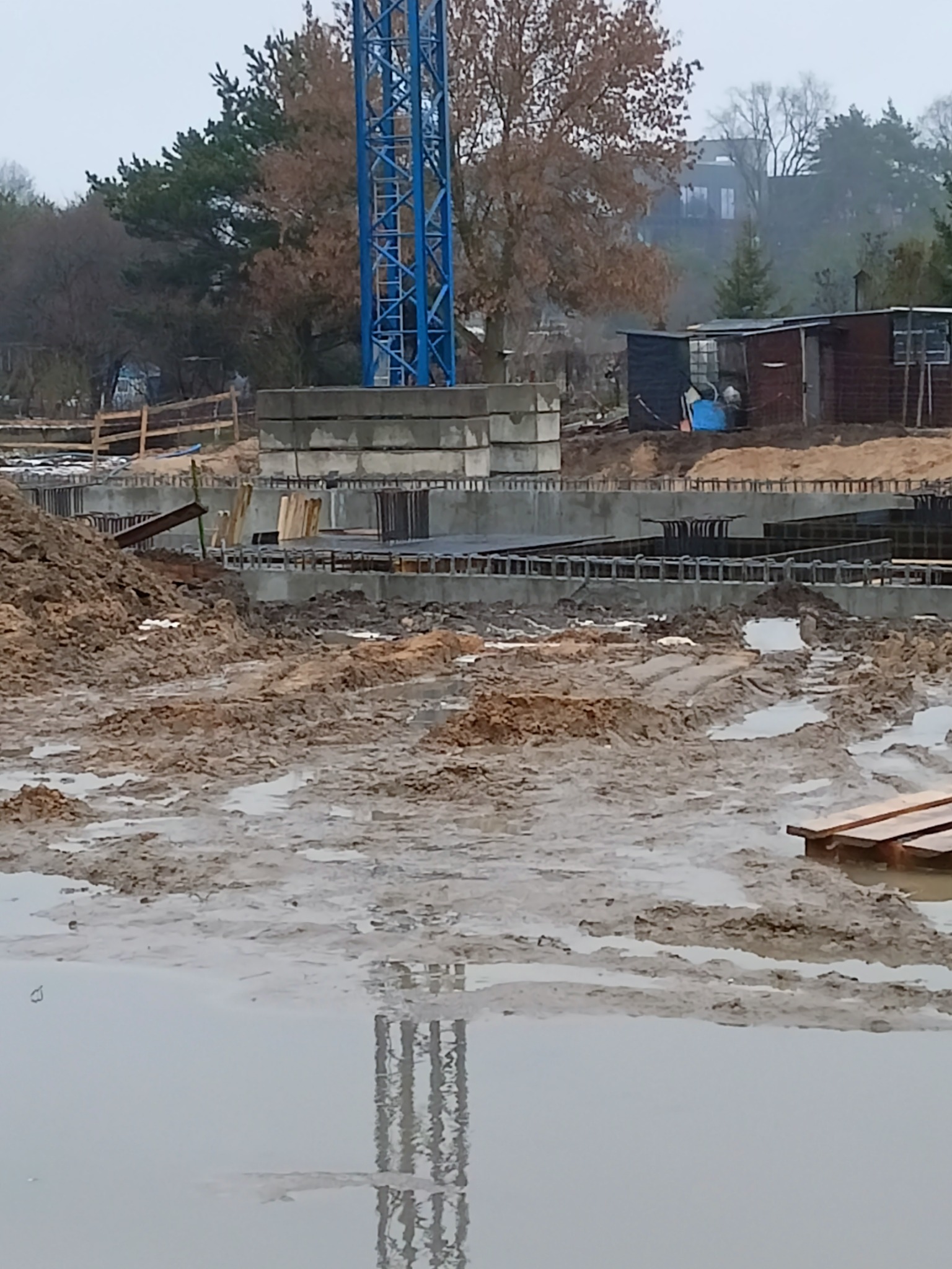 Fundamenty budynku w trakcie budowy, widoczne zbrojenia i betonowe elementy konstrukcyjne, w tle żuraw budowlany i drzewa, teren podmokły.