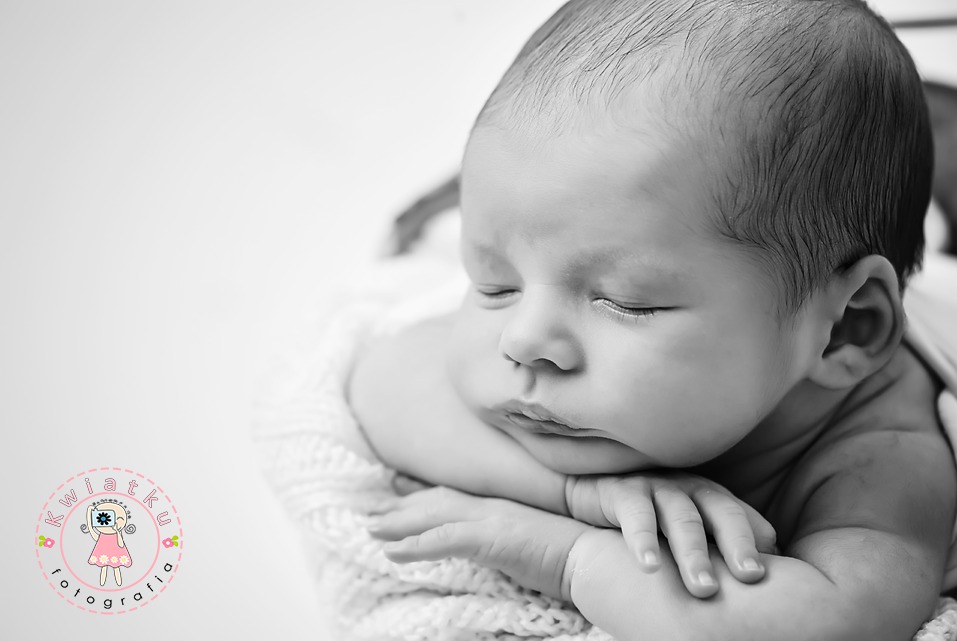 Czarno-białe, zbliżenie śpiącego noworodka z ciemnymi włoskami, leżącego na miękkim, białym kocyku, z rączkami pod brodą. Delikatne światło podkreśla rysy twarzy.