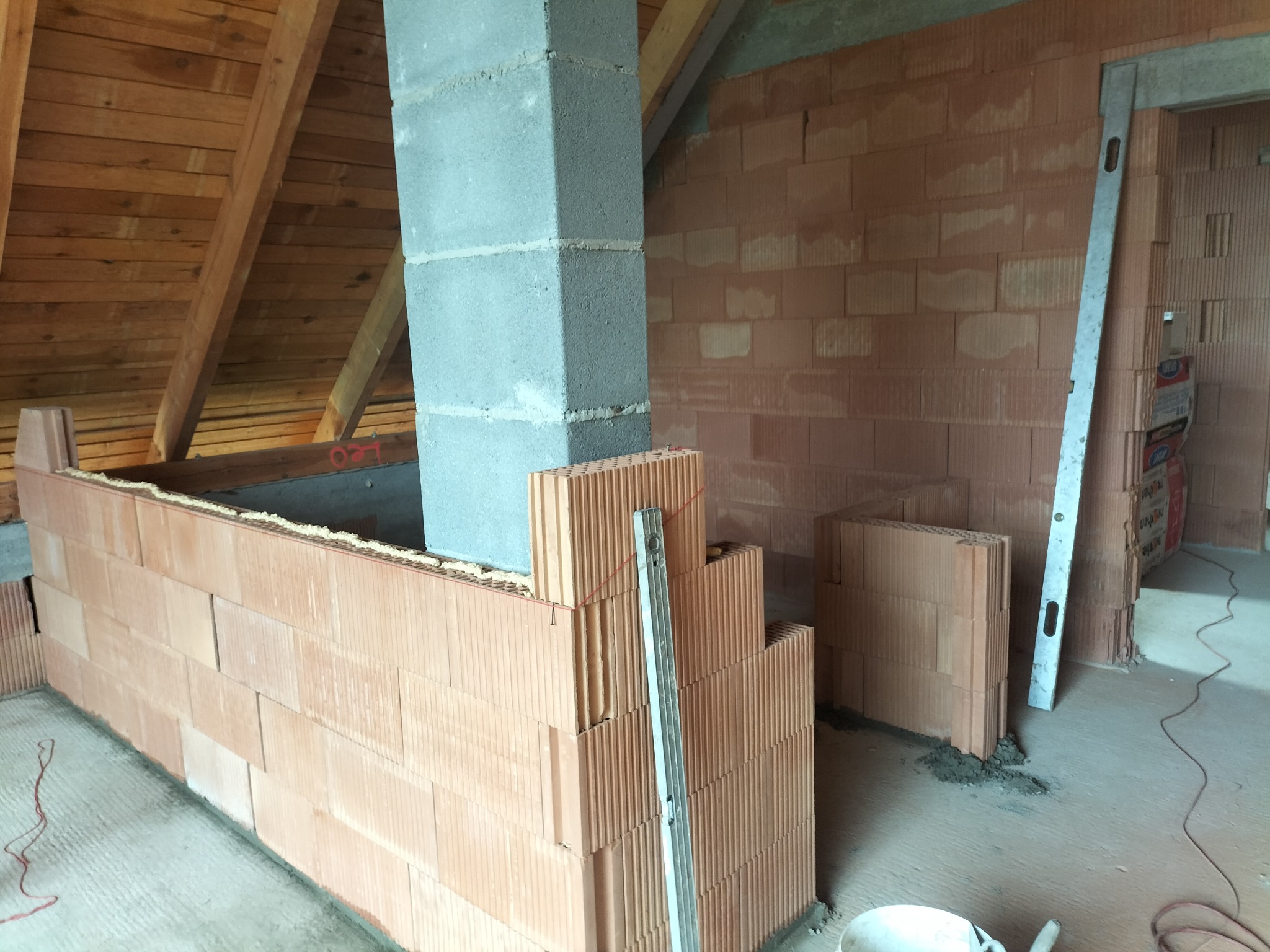 Wnętrze poddasza w trakcie murowania ścian z czerwonej cegły, z widocznym kominem z szarego betonu i poziomnicą opartą o ścianę. Etap budowy.