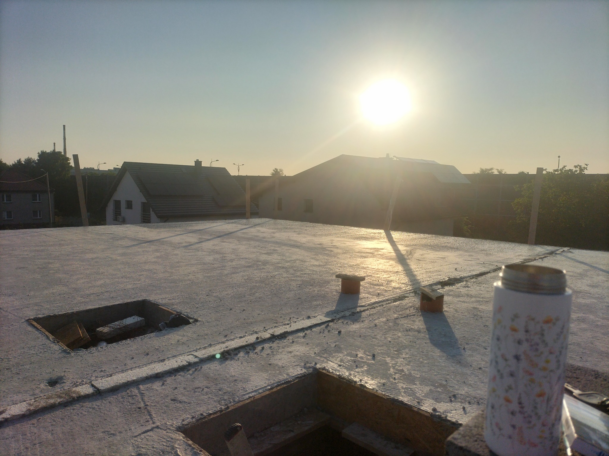 Świeżo wylany betonowy strop z otworami na instalacje, widok w kierunku zachodzącego słońca. W tle zabudowa jednorodzinna.