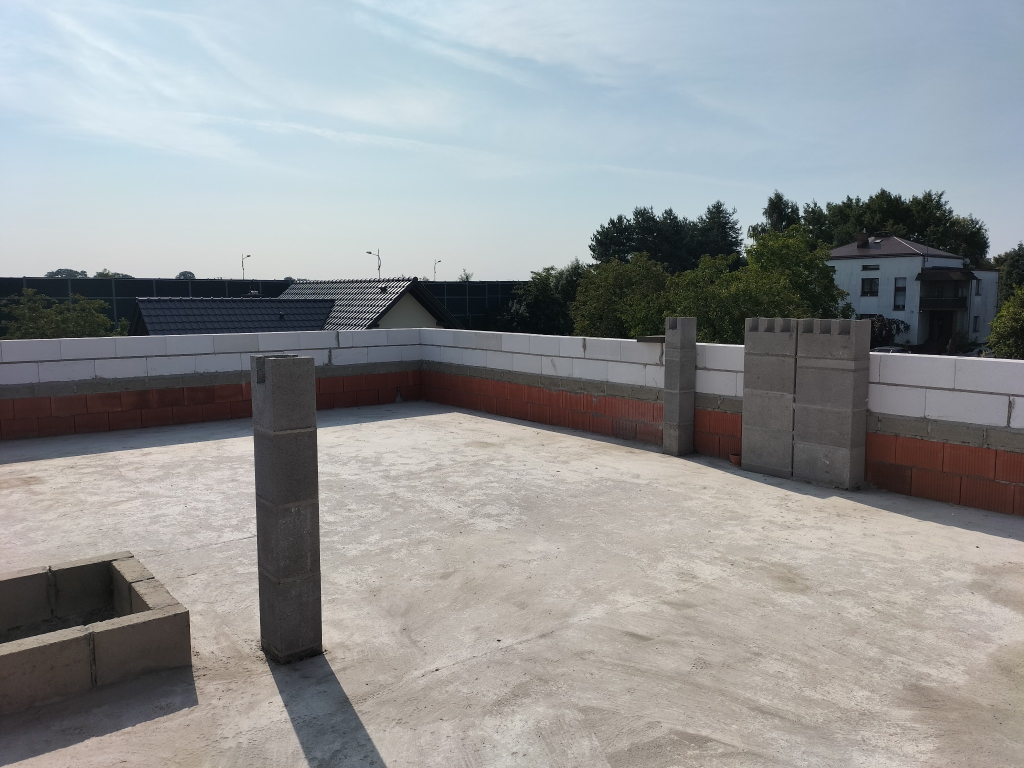 Wznoszenie ścian domu z pustaków i cegły, widok z góry na plac budowy w słoneczny dzień. Częściowo wymurowane ściany i filary. W tle domy i drzewa.