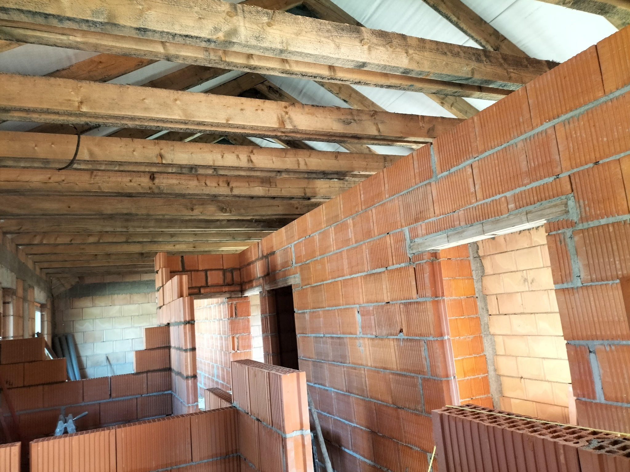 Wnętrze budynku w trakcie budowy. Widoczne ściany z czerwonej cegły, drewniana konstrukcja dachu i pustaki. Częściowo wymurowane ściany działowe.