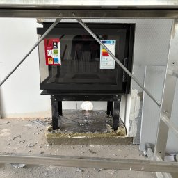 WOOD & STEEL HOME - Montaż wkładu kominkowego w surowym wnętrzu. Widoczny wkład z szybą, obudowa z izolacją i fragment rusztowania. Prace wykończeniowe w toku.