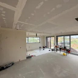 Wnętrze w trakcie prac wykończeniowych: płyty karton-gips na suficie, szpachlowane łączenia, surowa podłoga, duże okna z widokiem na zewnątrz. Widoczne narzędzia i materiały.