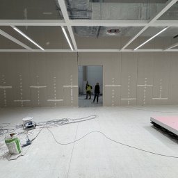 WOOD & STEEL HOME - Montaż płyt karton-gips na ścianach w dużym pomieszczeniu. Widoczne profile sufitowe, narzędzia i materiały budowlane. Dwóch pracowników w oddali.