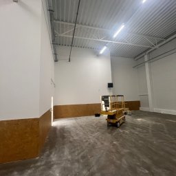 WOOD & STEEL HOME - Wnętrze hali przemysłowej po remoncie, z podnośnikiem nożycowym na środku. Ściany pomalowane na biało, z brązowym pasem na dole. Widoczna instalacja pod sufitem.