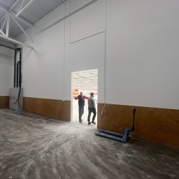 WOOD & STEEL HOME - Wnętrze hali magazynowej z surową podłogą, białą ścianą z brązowym panelem i dwoma osobami w oddali. W rogu wózek paletowy.