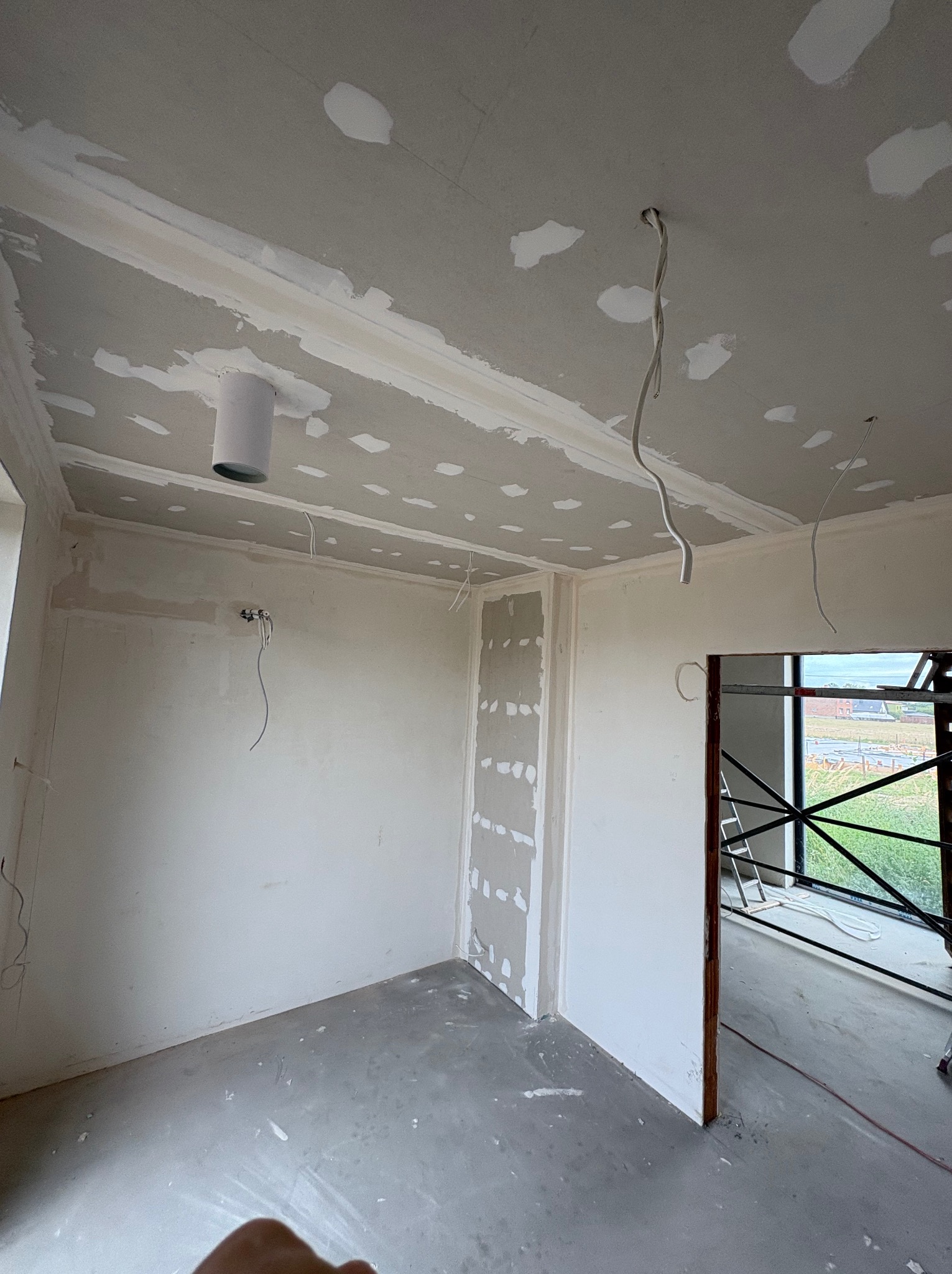 Wnętrze w trakcie remontu: surowy sufit z widocznymi punktowymi poprawkami gładzi, wystające przewody elektryczne i szara, surowa podłoga. W tle widoczne rusztowanie za oknem.
