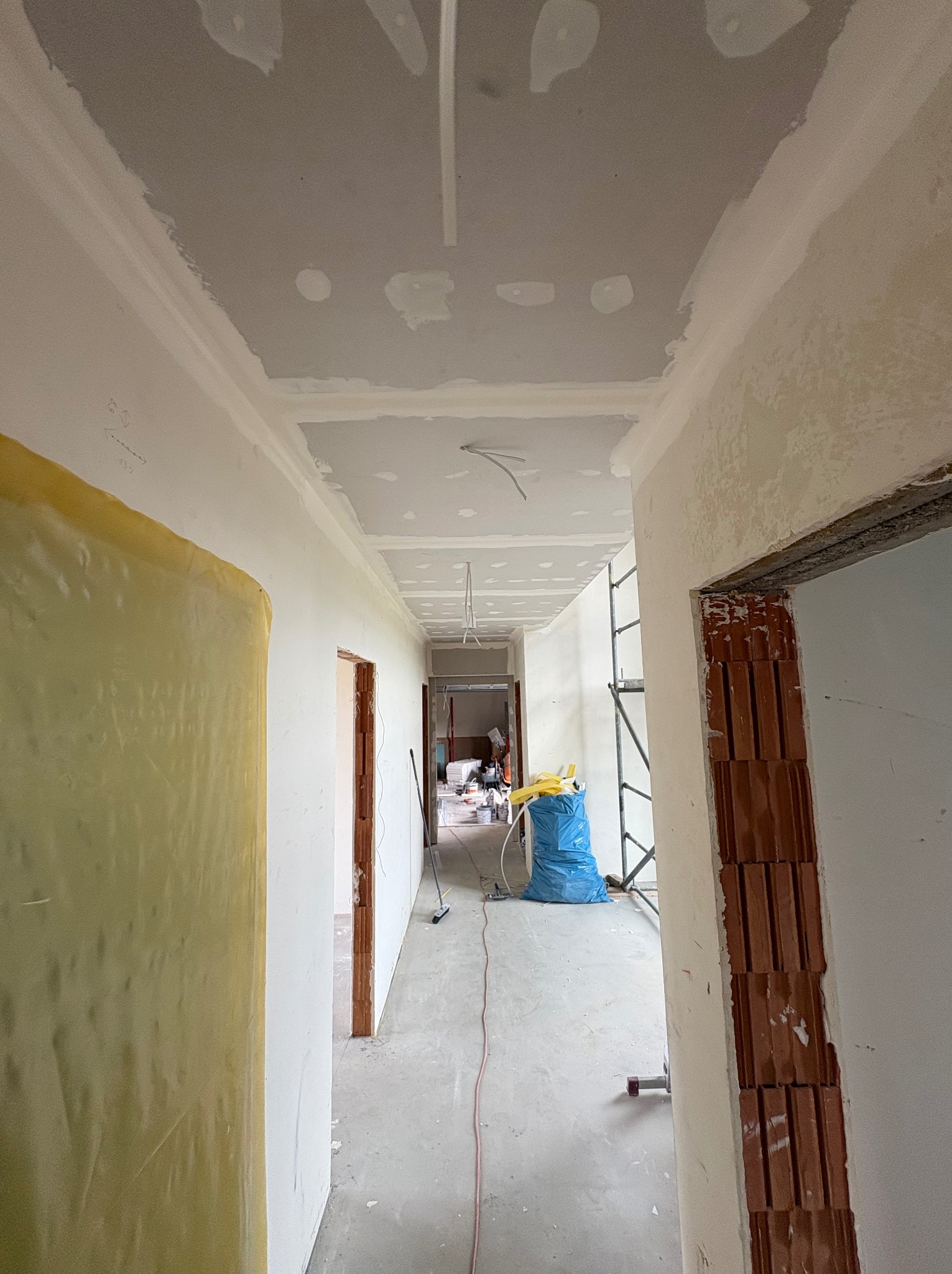 Korytarz w trakcie prac wykończeniowych: widoczne płyty karton-gips na suficie, przygotowane ściany, zabezpieczona folią ściana i gruz w worku. Prace remontowe.