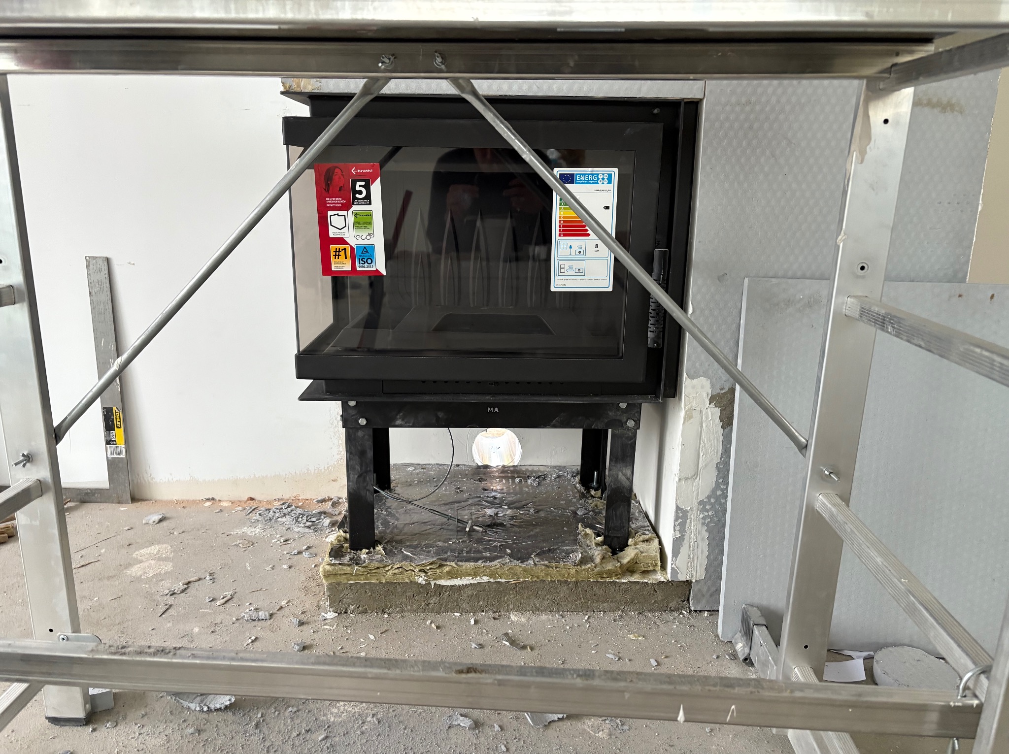 Montaż wkładu kominkowego w surowym wnętrzu. Widoczny wkład z szybą, obudowa z izolacją i fragment rusztowania. Prace wykończeniowe w toku.