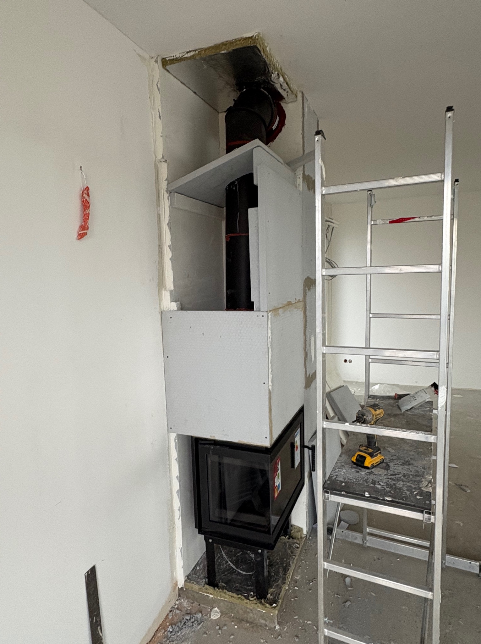 Wnętrze w trakcie zabudowy kominka płytami gipsowo-kartonowymi. Widoczna drabina, narzędzia i fragment instalacji kominowej. Prace wykończeniowe.
