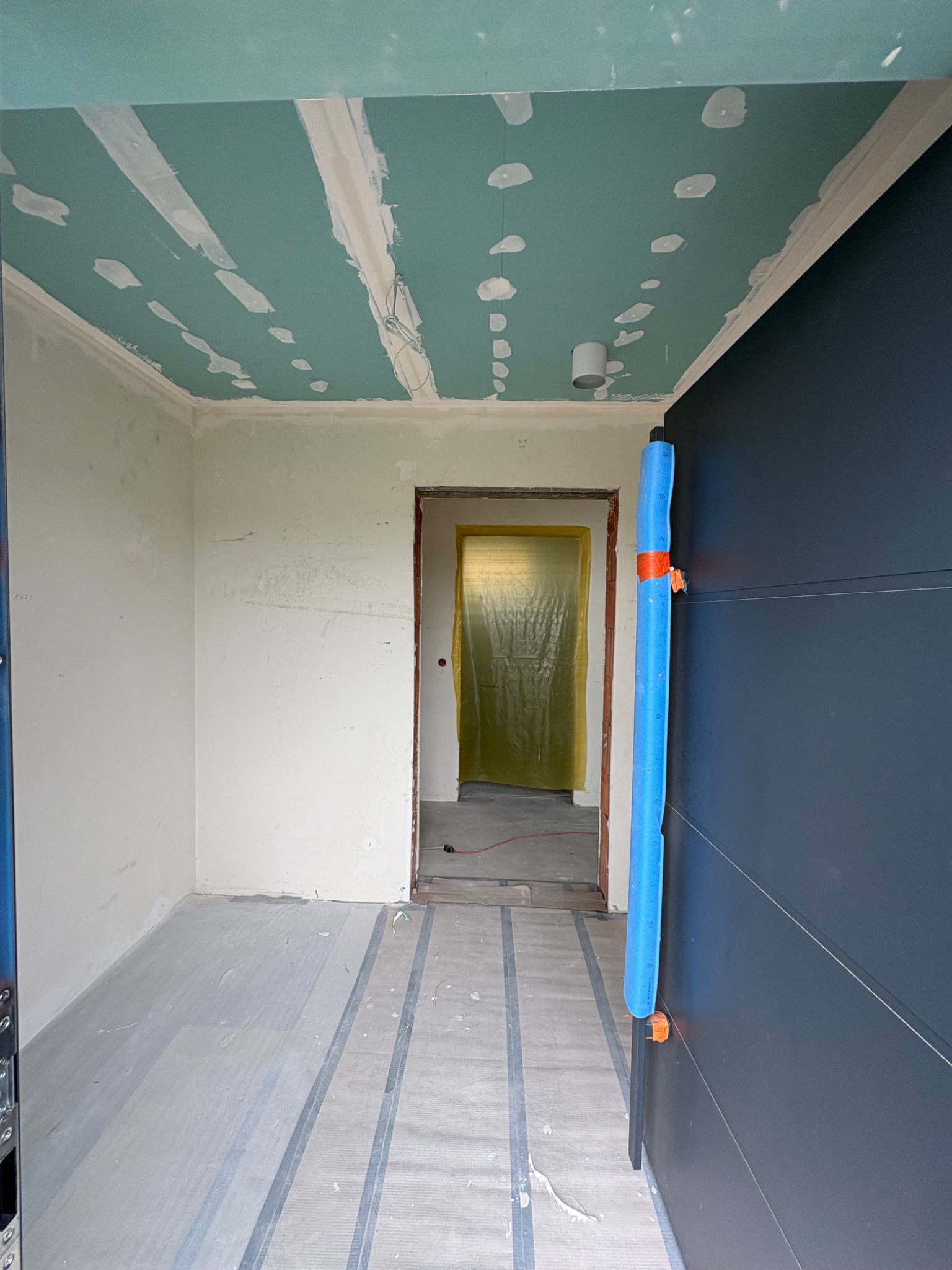Wnętrze w trakcie remontu: sufit z płyt karton-gips, częściowo zaszpachlowany, zabezpieczona podłoga, widok na kolejne pomieszczenie z folią ochronną.