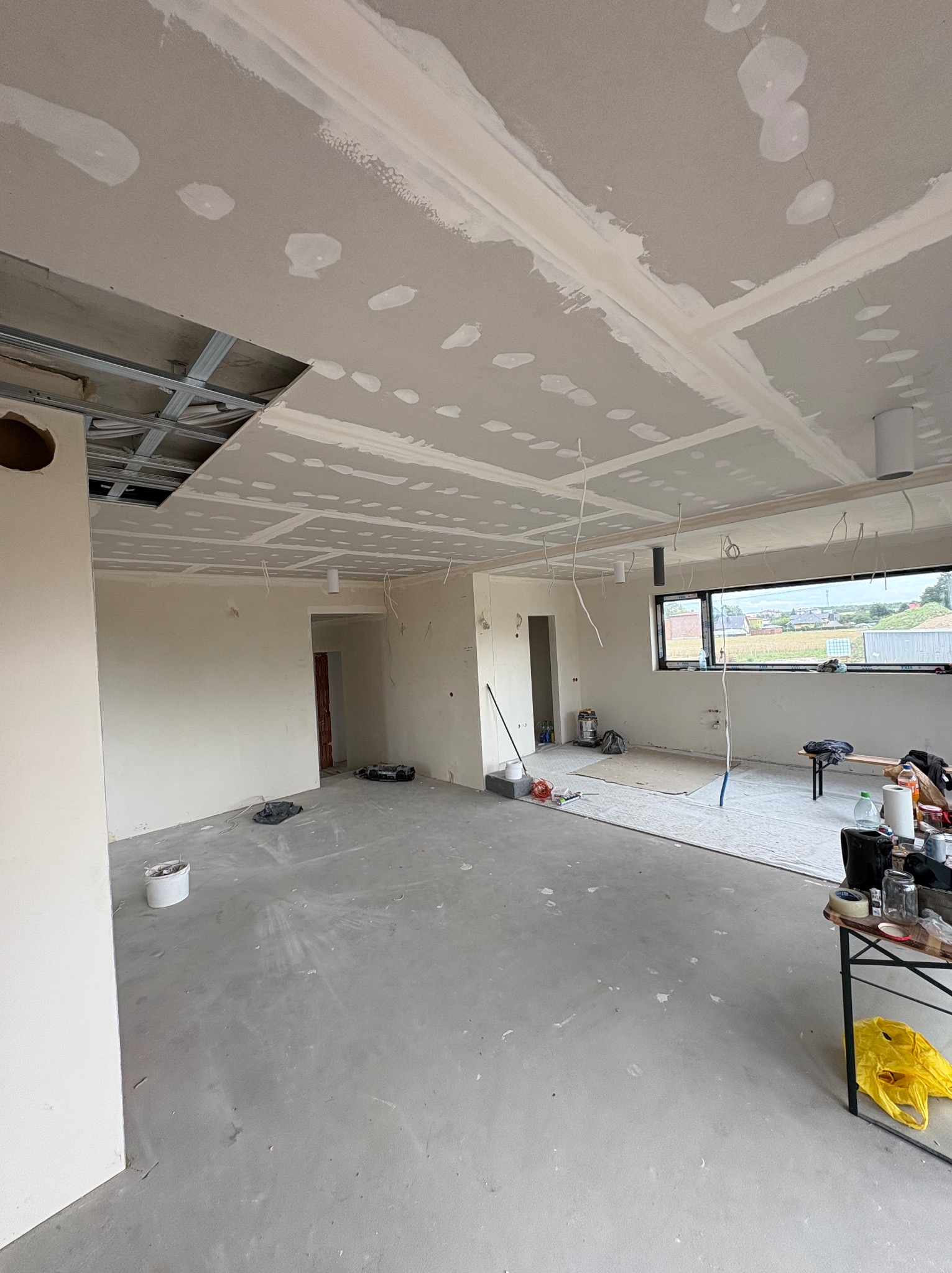 Wnętrze w trakcie montażu płyt karton-gips na suficie, widoczne metalowe profile, kable, szpachlowane łączenia. Częściowo wykończone ściany i surowy beton na podłodze.