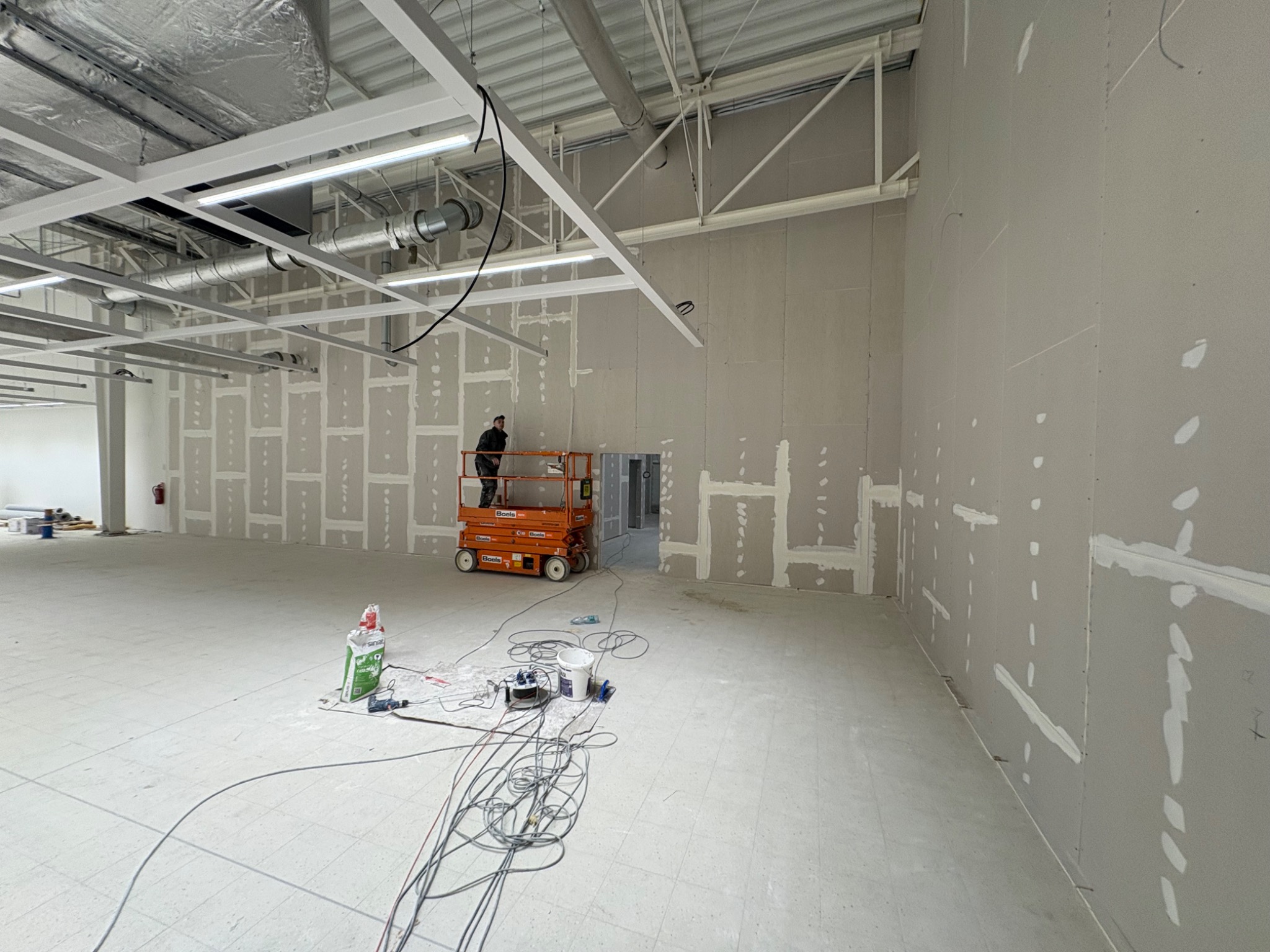 Montaż płyt karton-gips w dużym pomieszczeniu. Widoczny pracownik na podnośniku, ściany z częściowo zamontowanymi płytami i narzędzia pracy na podłodze.