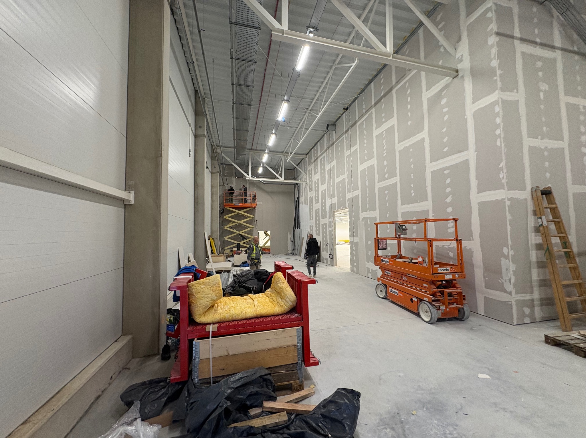 Montaż płyt karton-gips w hali magazynowej. Widoczne podnośniki, rusztowania i częściowo zamontowane płyty na ścianach. Prace wykończeniowe w surowym wnętrzu.