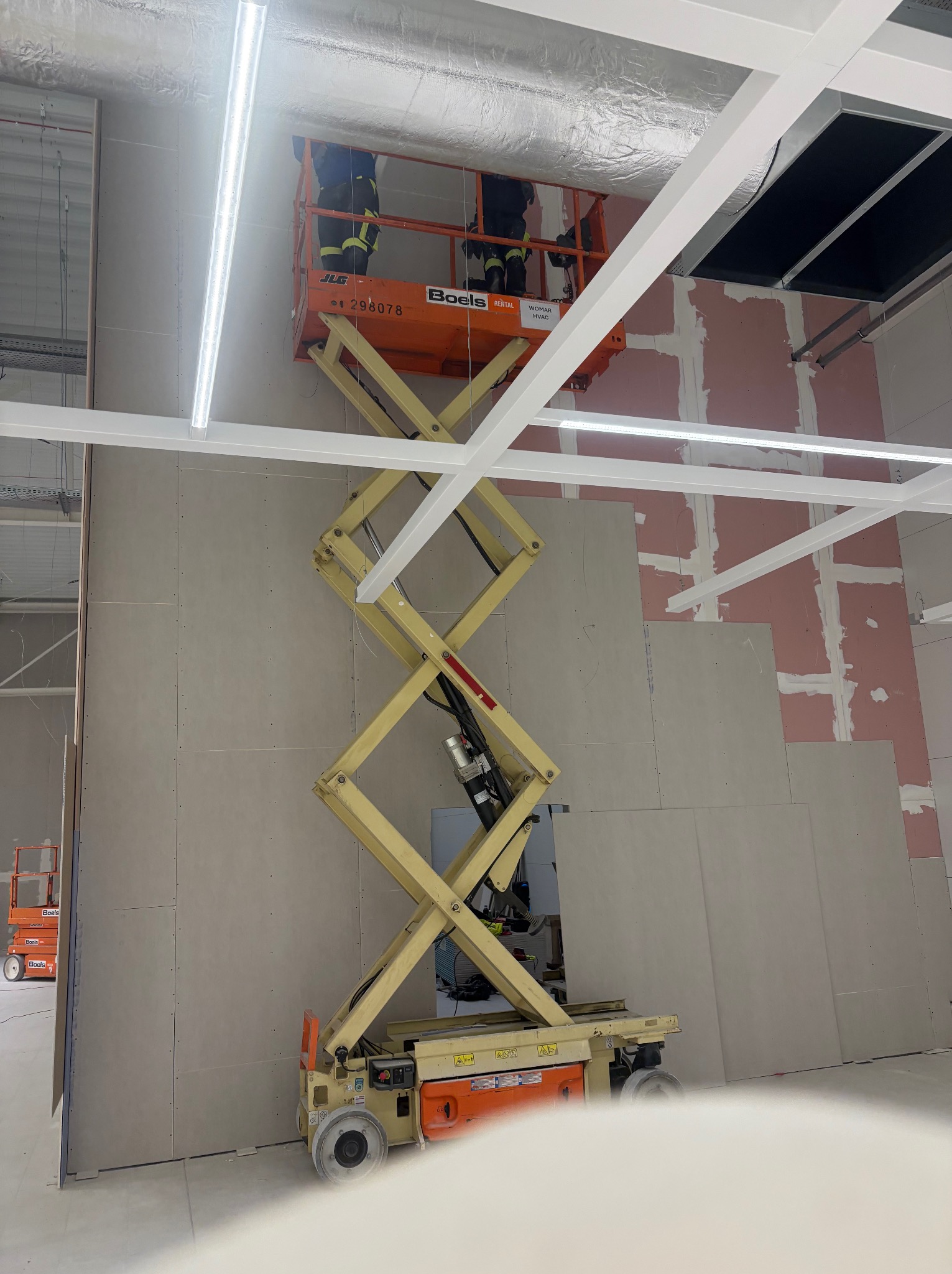 Montaż płyt karton-gips na ścianie w dużym pomieszczeniu z użyciem podnośnika nożycowego. Widoczne profile sufitowe i częściowo wykończona ściana.