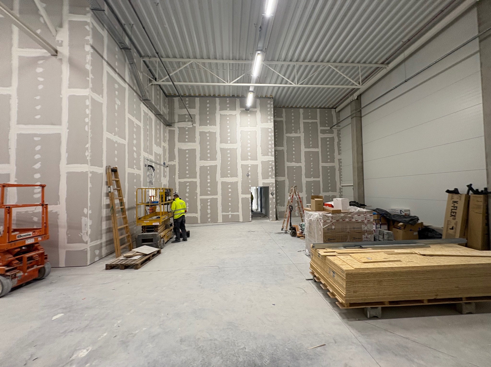 Montaż płyt karton-gips w dużym pomieszczeniu. Widoczne rusztowania, materiały budowlane i pracownik w odblaskowej kamizelce. Ściany pokryte płytami, przygotowane do dalszych prac.