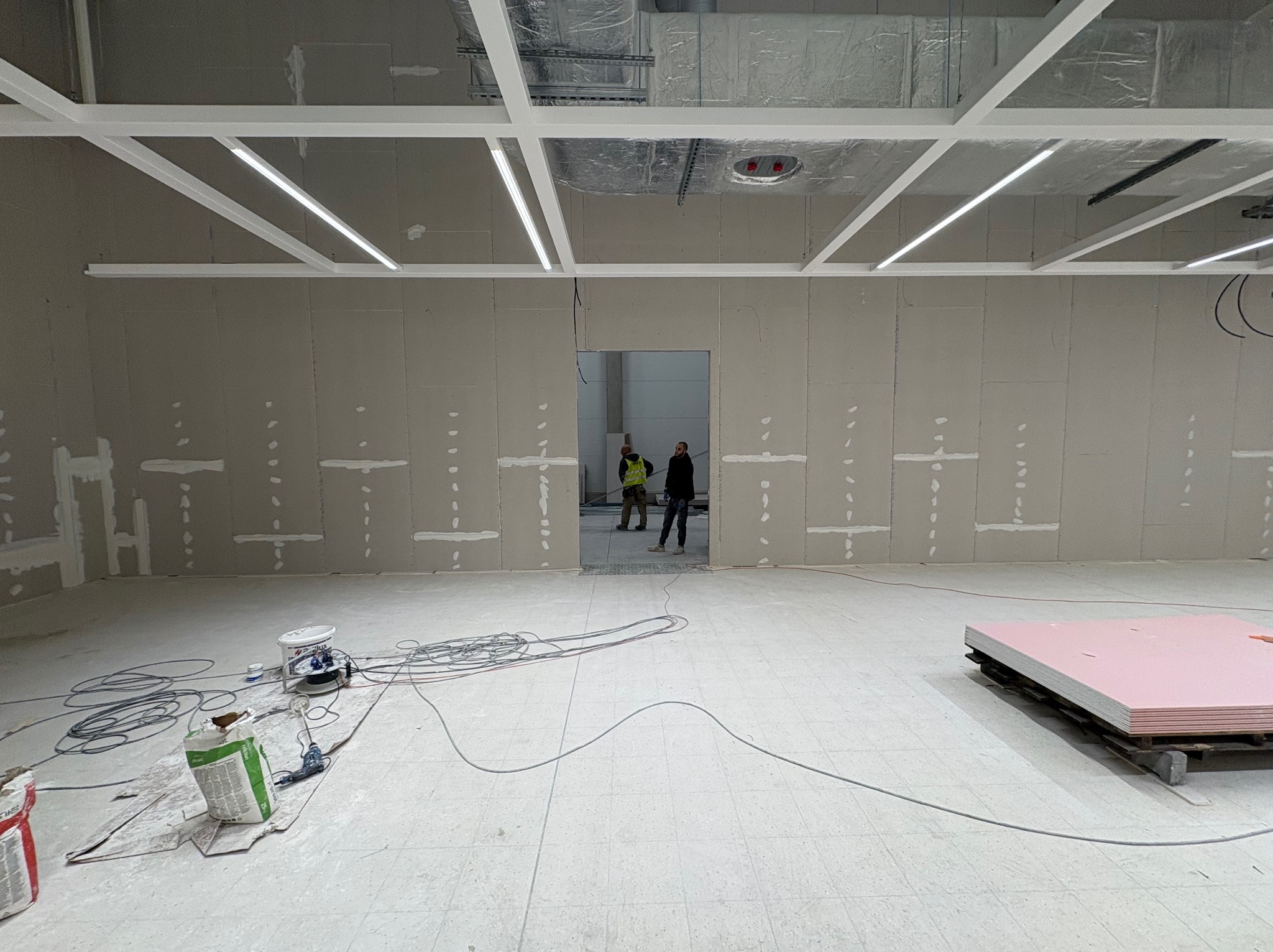 Montaż płyt karton-gips na ścianach w dużym pomieszczeniu. Widoczne profile sufitowe, narzędzia i materiały budowlane. Dwóch pracowników w oddali.