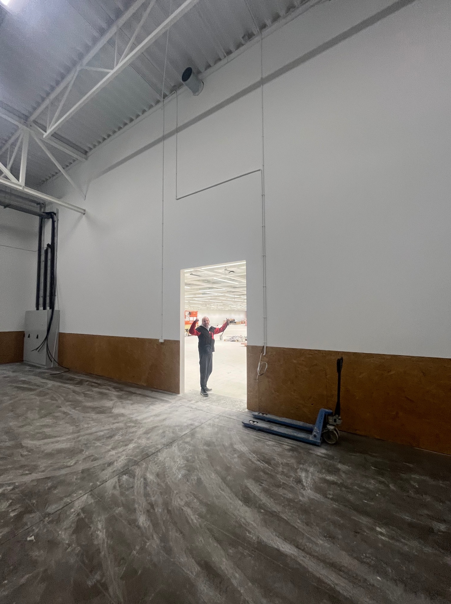 Wnętrze hali w trakcie prac wykończeniowych: gładkie, białe ściany z brązowym pasem, betonowa posadzka z widocznymi śladami, w tle pracownik z podniesionymi rękami.