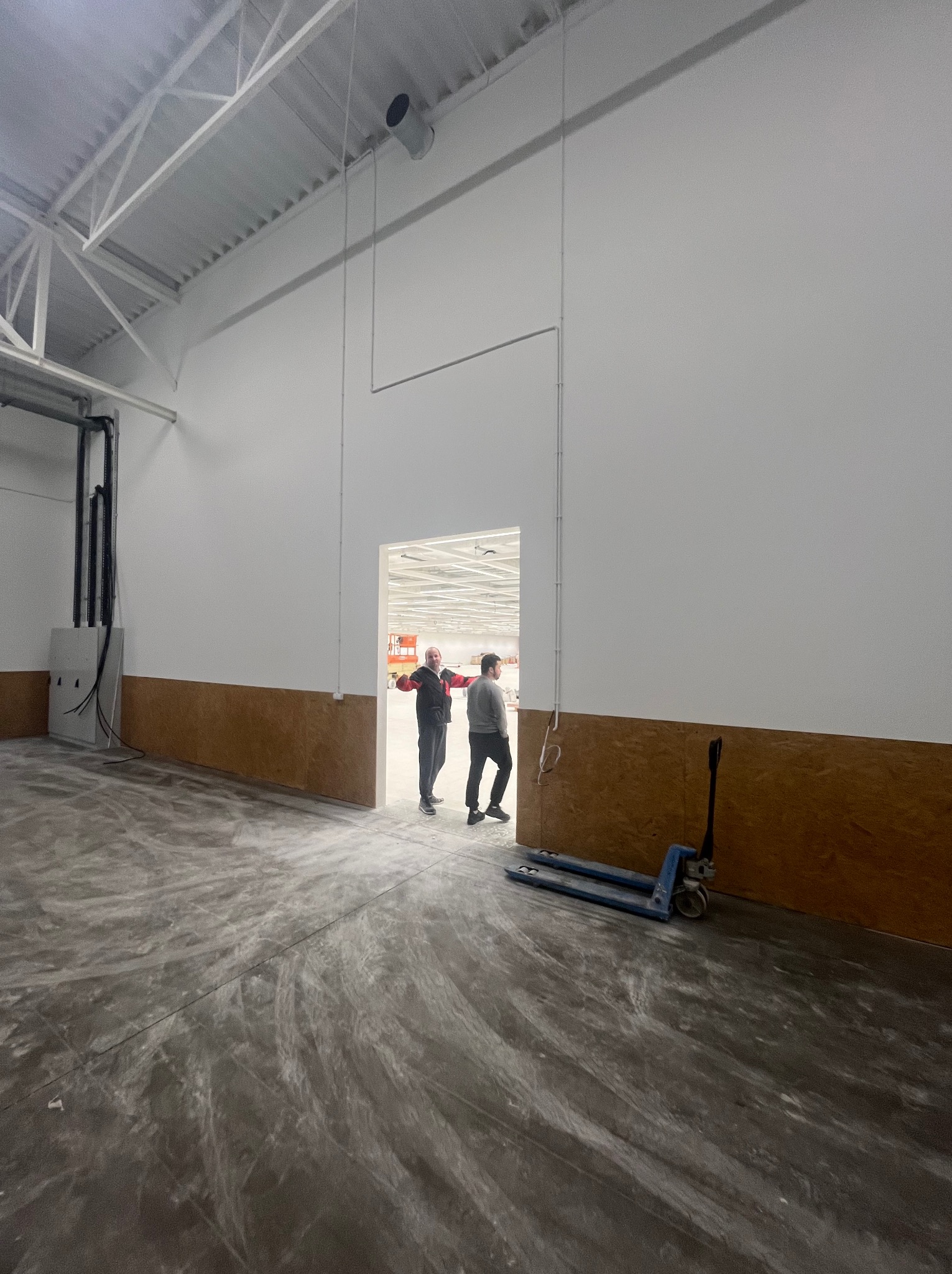 Wnętrze hali magazynowej z surową podłogą, białą ścianą z brązowym panelem i dwoma osobami w oddali. W rogu wózek paletowy.