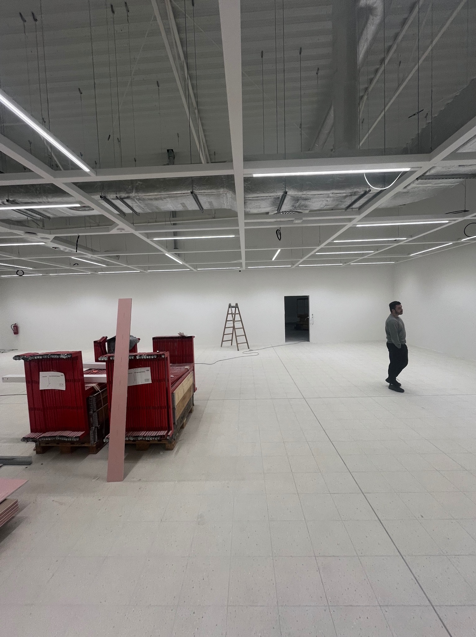 Wnętrze w trakcie prac wykończeniowych: widoczne materiały budowlane, drabina, instalacja pod sufitem i mężczyzna w oddali. Duża przestrzeń z białymi ścianami i podłogą.