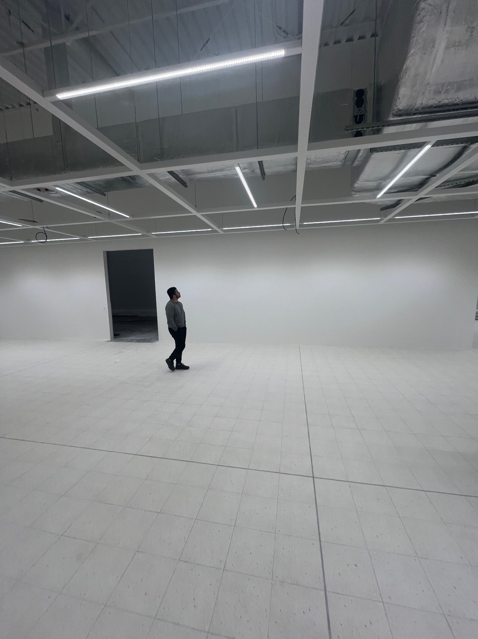 Wnętrze w trakcie wykończenia: białe ściany, podłoga z płytek, sufit z instalacją oświetleniową. Mężczyzna w oddali, w szarym ubraniu, patrzy w górę. Widoczny otwór drzwiowy.