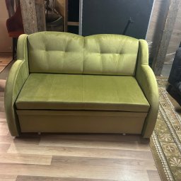 Zakład tapicerski VIOLA - Sofa dwuosobowa w odcieniach zieleni, z pikowanymi poduchami oparcia, na tle drewnianej podłogi i elementów konstrukcyjnych budynku.