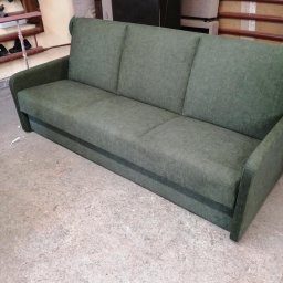 Zakład tapicerski VIOLA - Zielona, trzyosobowa sofa z podłokietnikami, stoi na betonowej podłodze. W tle widoczne elementy warsztatu tapicerskiego i inne meble.