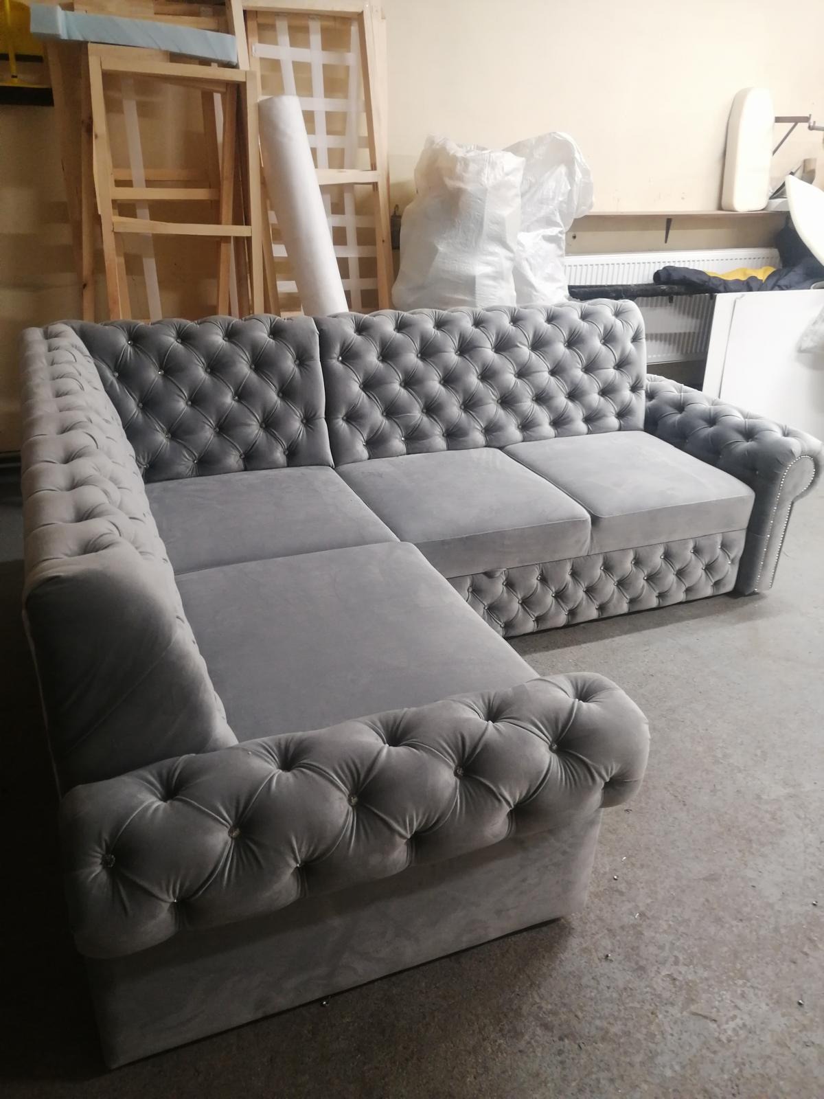 Szara, tapicerowana sofa narożna typu chesterfield z guzikami, widoczna w warsztacie tapicerskim. W tle ramy i materiały tapicerskie.