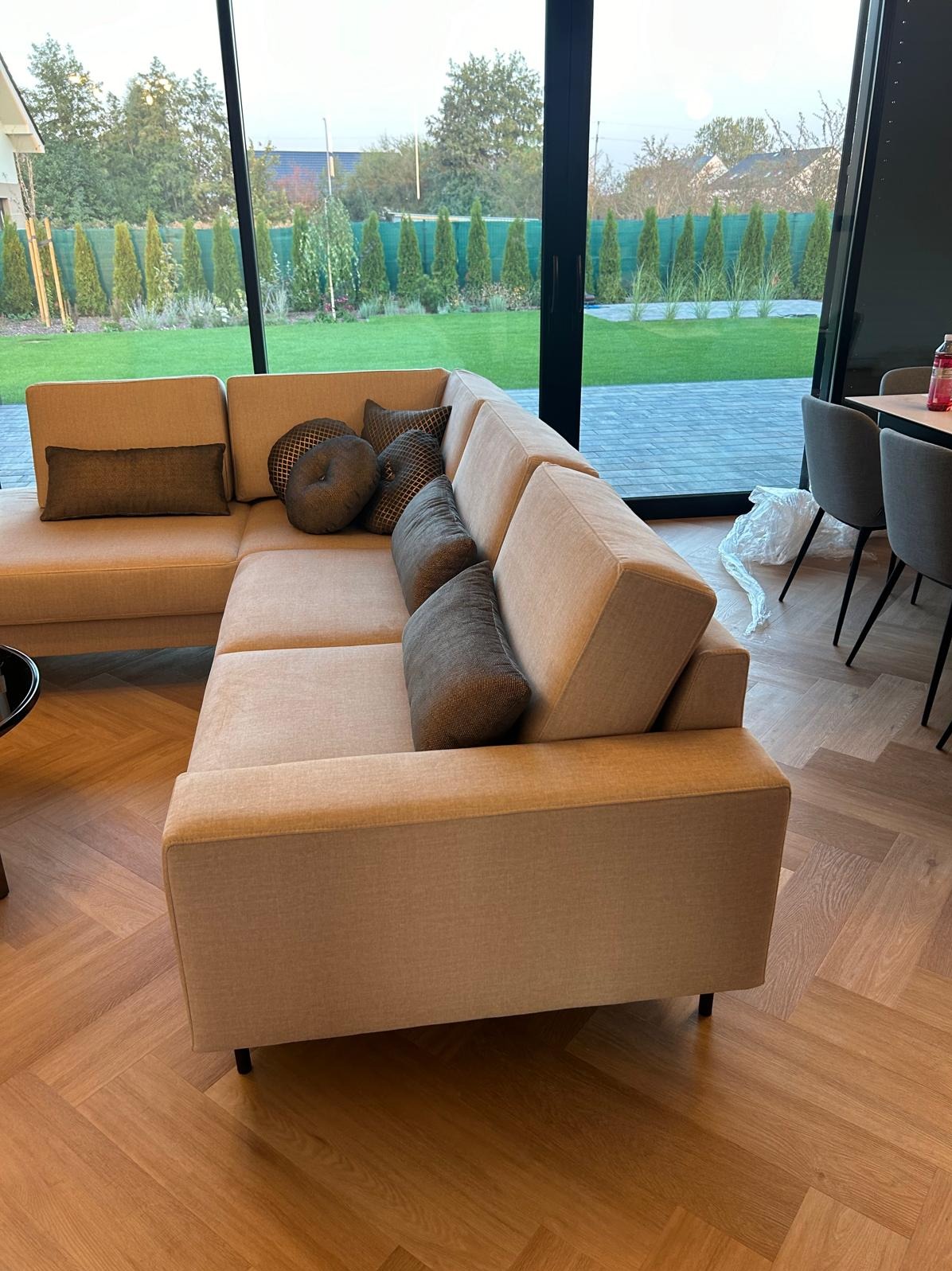 Beżowa sofa narożna z poduszkami w nowoczesnym salonie, widok na ogród przez panoramiczne okno. Parkiet w jodełkę, minimalistyczny design.