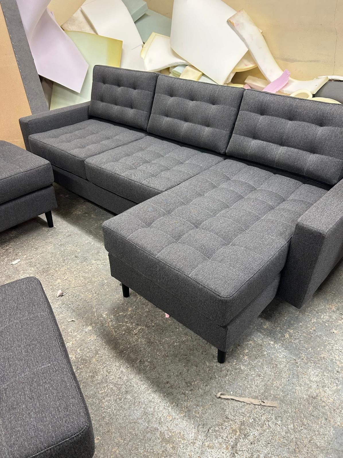 Szara, narożna sofa tapicerowana z pikowanymi siedziskami i poduszkami, uzupełniona o dwa pufy. W tle widoczne materiały tapicerskie.