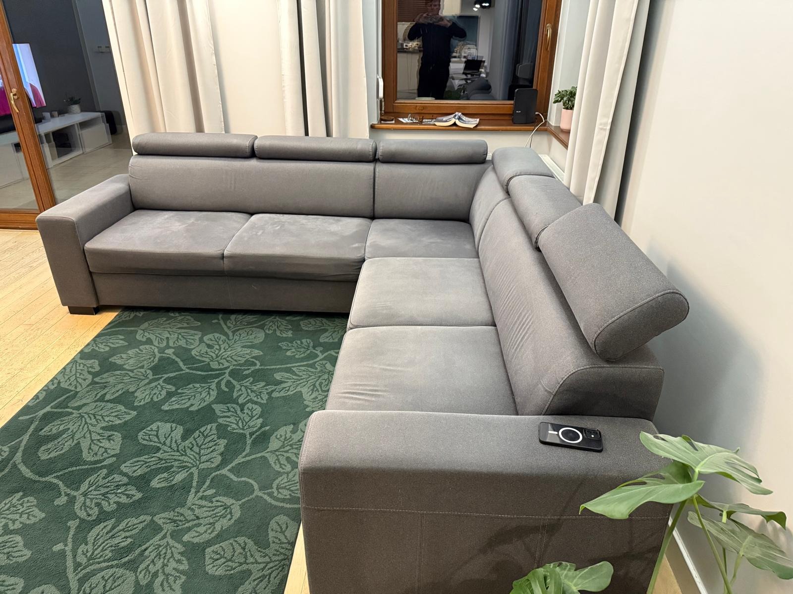 Szara, narożna kanapa z regulowanymi zagłówkami, ustawiona na zielonym dywanie w salonie. Na podłokietniku leży smartfon. Widoczne okno z odbiciem osoby.