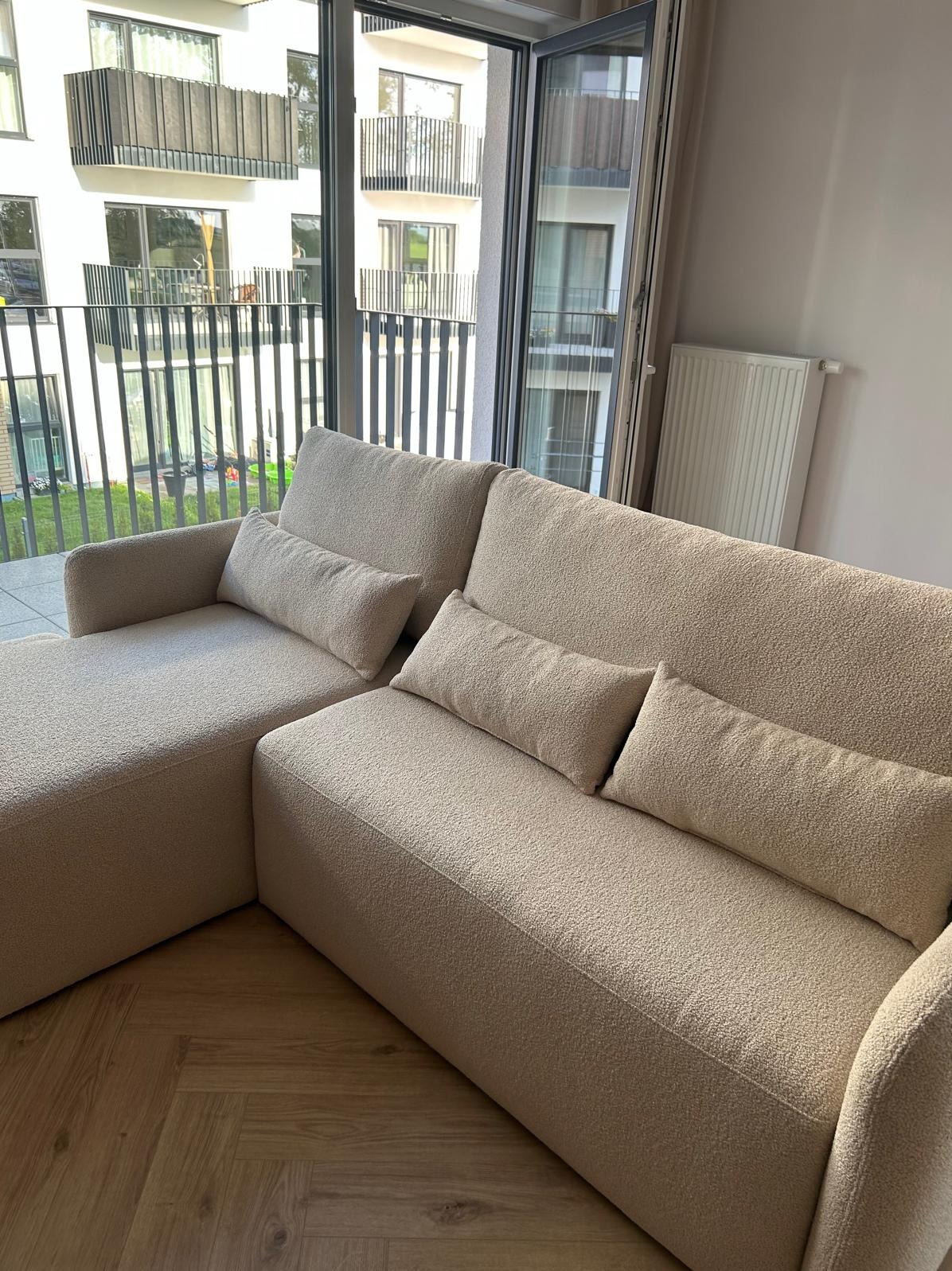 Beżowa, modułowa sofa z poduszkami w nowoczesnym salonie, widok na balkon i budynek mieszkalny zza okna. Parkiet na podłodze.