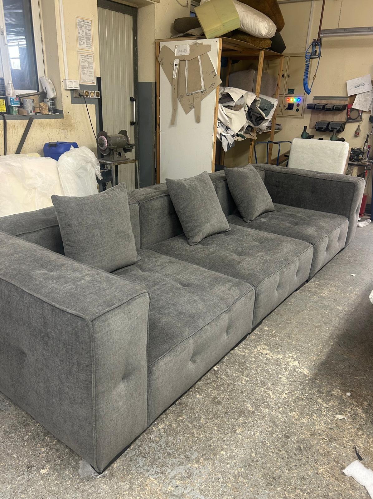 Szara, modułowa sofa z poduszkami w warsztacie tapicerskim. Widoczne narzędzia i materiały, co sugeruje etap produkcji lub renowacji.