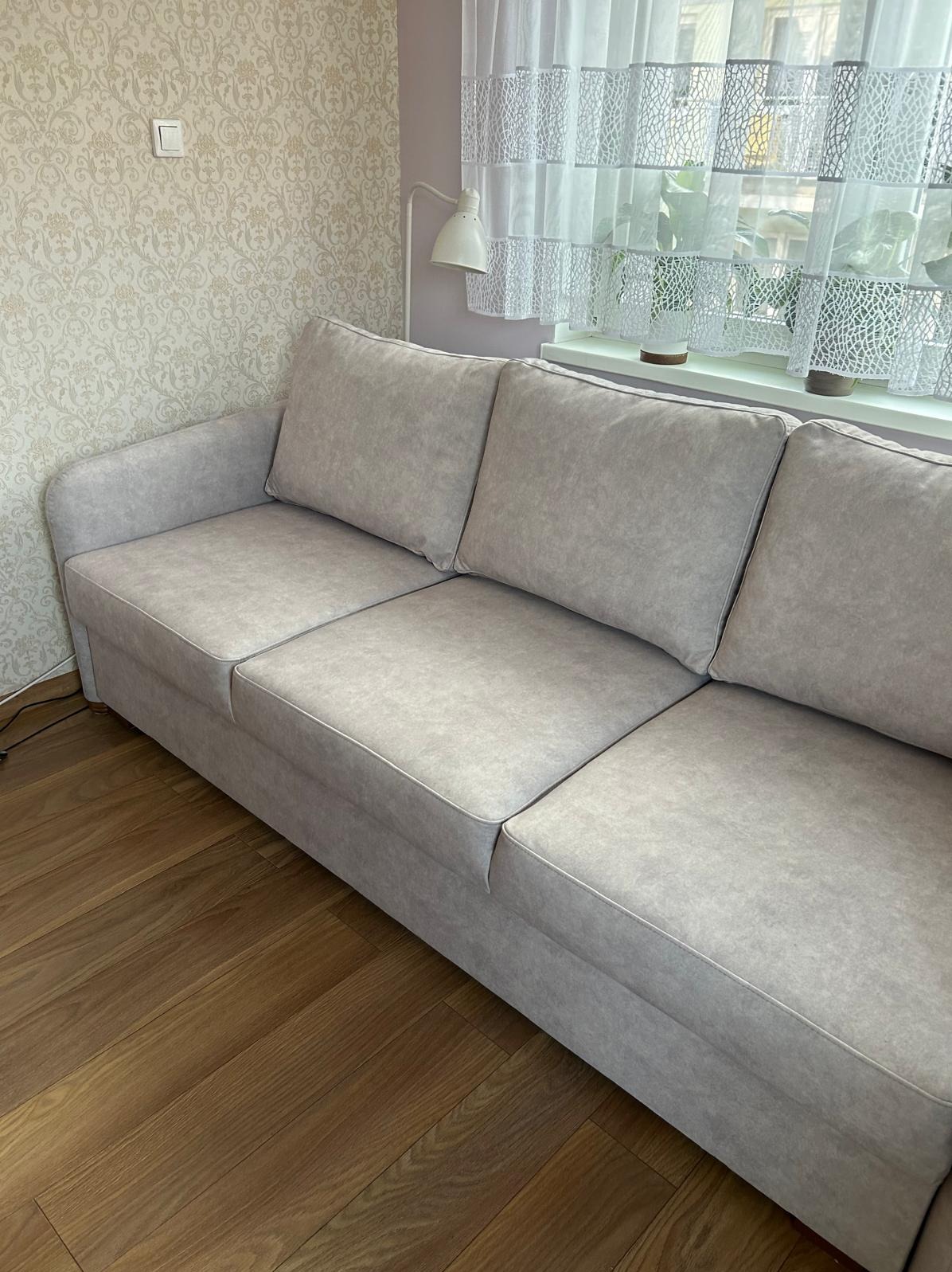 Jasnoszara, tapicerowana sofa z trzema poduszkami na drewnianej podłodze, w tle okno z białymi zasłonami i wzorzysta tapeta. Stonowana kolorystyka.