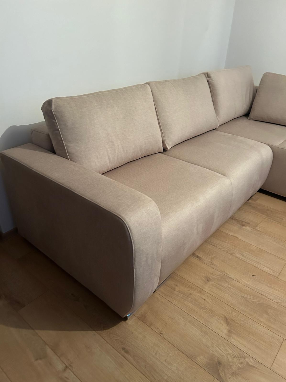 Beżowy narożnik z tapicerowanym obiciem, widoczny z boku na tle parkietu i białej ściany. Minimalistyczny design, gładka tkanina, nowoczesny styl.