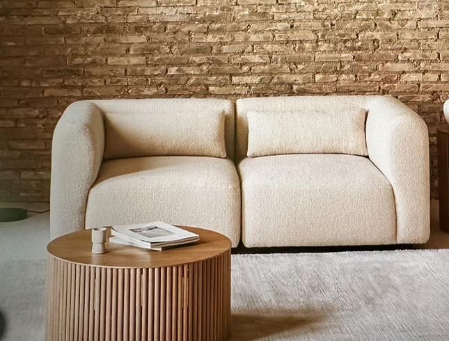 Beżowa, modułowa sofa z poduszką, obok okrągły stolik z książką, na tle ściany z cegły. Minimalistyczny design, teksturowana tapicerka.