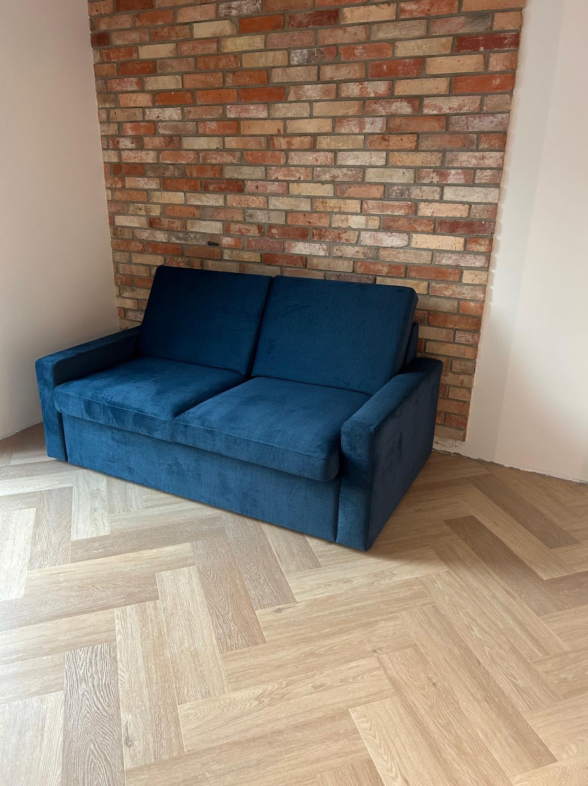 Niebieska sofa z aksamitną tapicerką na parkiecie w jodełkę, na tle ceglanej ściany. Minimalistyczny design, stonowana kolorystyka, nowoczesny styl.