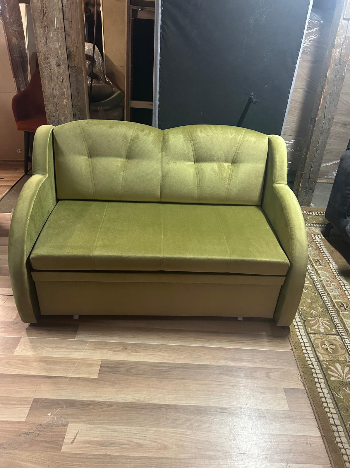 Sofa dwuosobowa w odcieniach zieleni, z pikowanymi poduchami oparcia, na tle drewnianej podłogi i elementów konstrukcyjnych budynku.