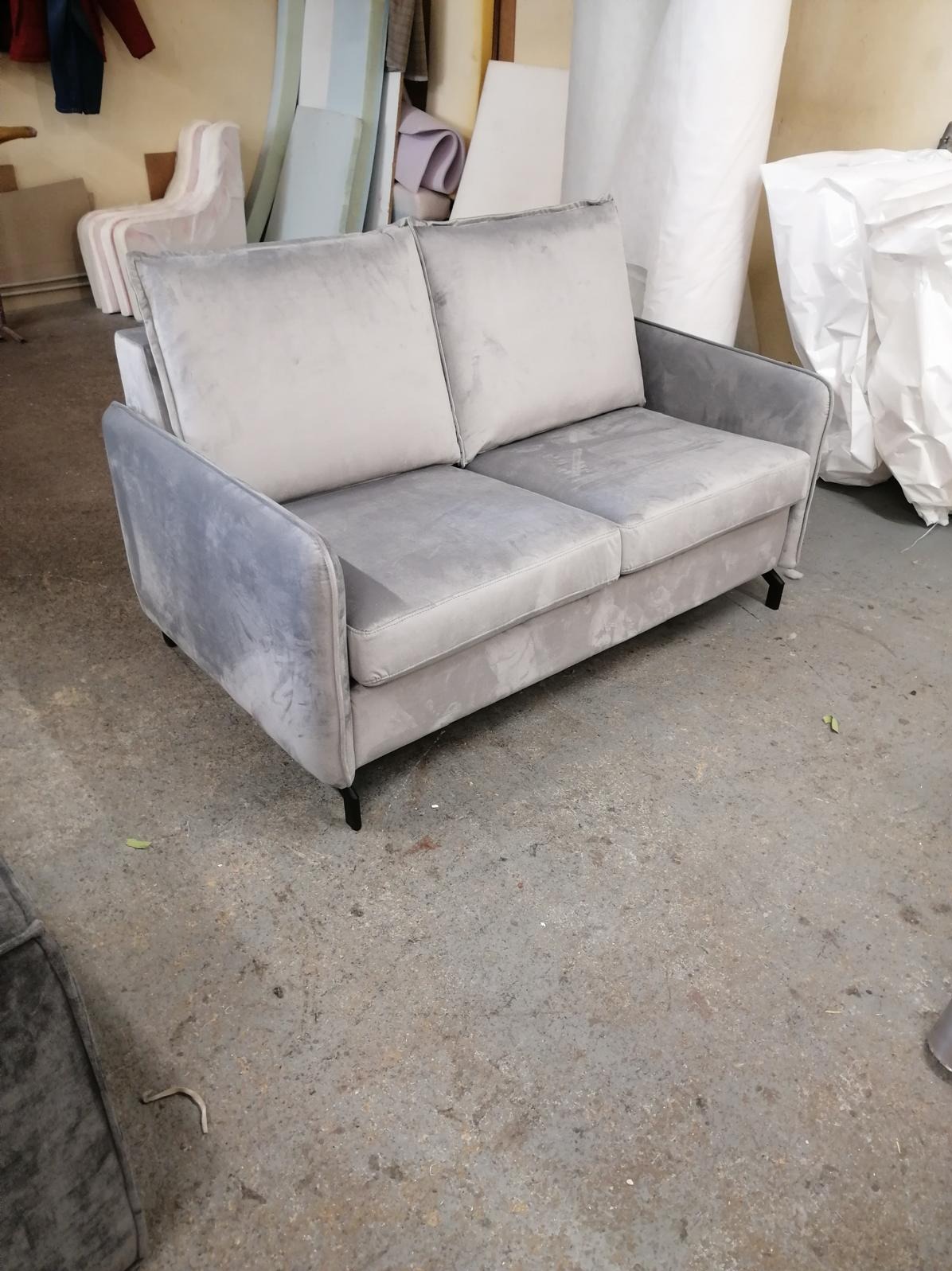 Dwuosobowa, szara sofa z weluru na niskich, czarnych nóżkach. W tle widoczne materiały tapicerskie i elementy mebli w warsztacie.