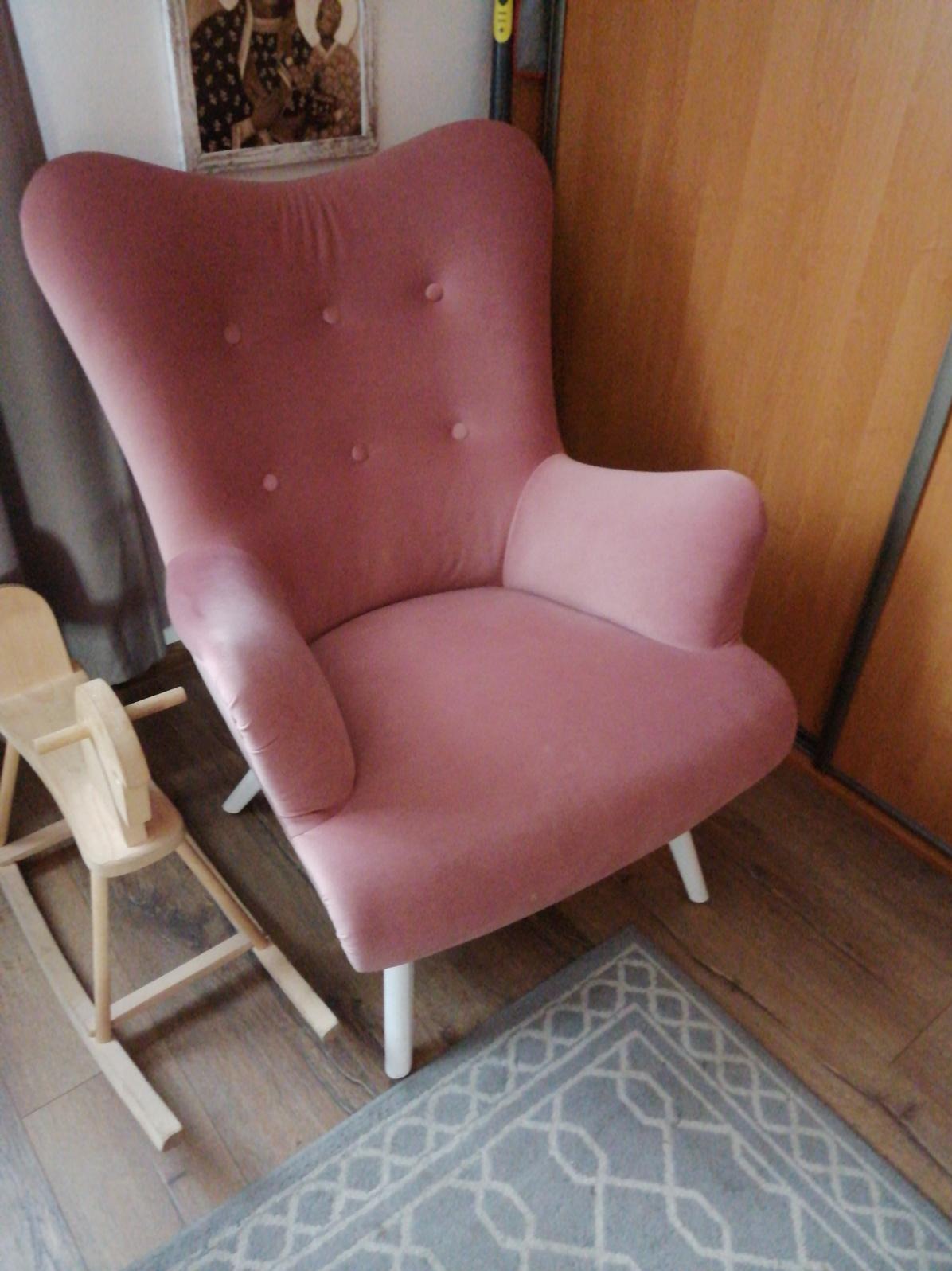 Różowy fotel uszak z guzikami na oparciu, drewniane nogi, obok drewniany konik na biegunach i dywan w geometryczne wzory, na tle drewnianych drzwi.