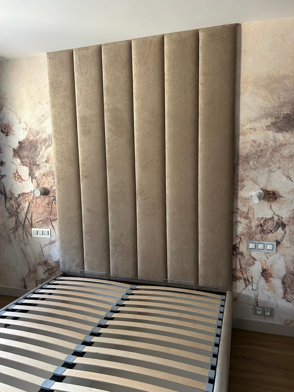 Beżowy, tapicerowany zagłówek łóżka z pionowymi panelami na tle ściany w kwiaty. Widoczny drewniany stelaż łóżka. Oświetlenie boczne.