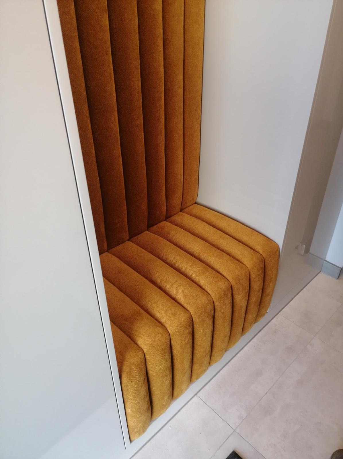 Zabudowany, pomarańczowy fotel tapicerowany z pionowymi przeszyciami, wkomponowany w białą wnękę. Widok z góry, ukazujący detale tkaniny i minimalistyczny design.