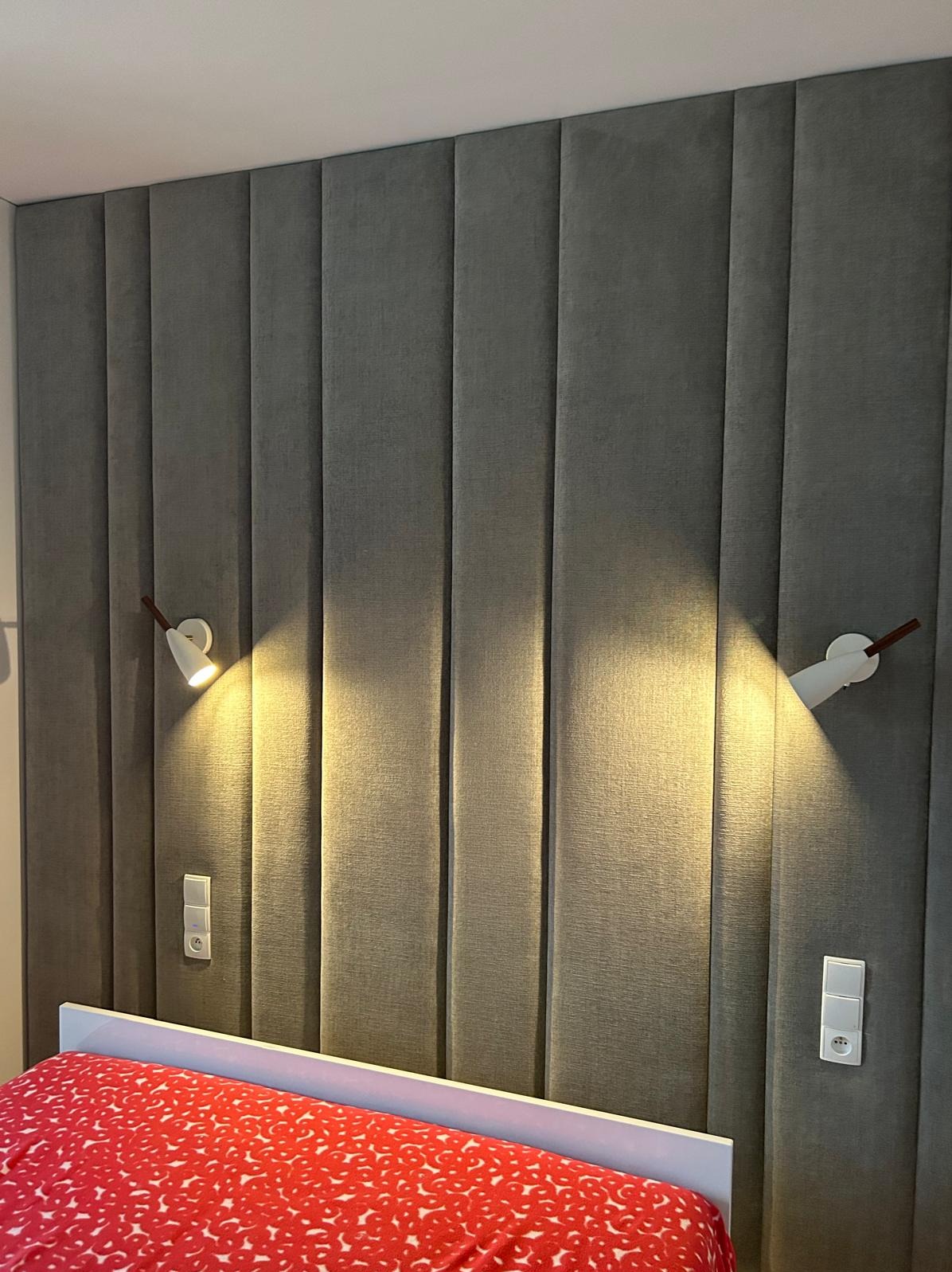 Tapicerowany panel ścienny w odcieniach szarości, oświetlony dwoma kinkietami. Fragment łóżka z czerwoną pościelą w dolnej części kadru.